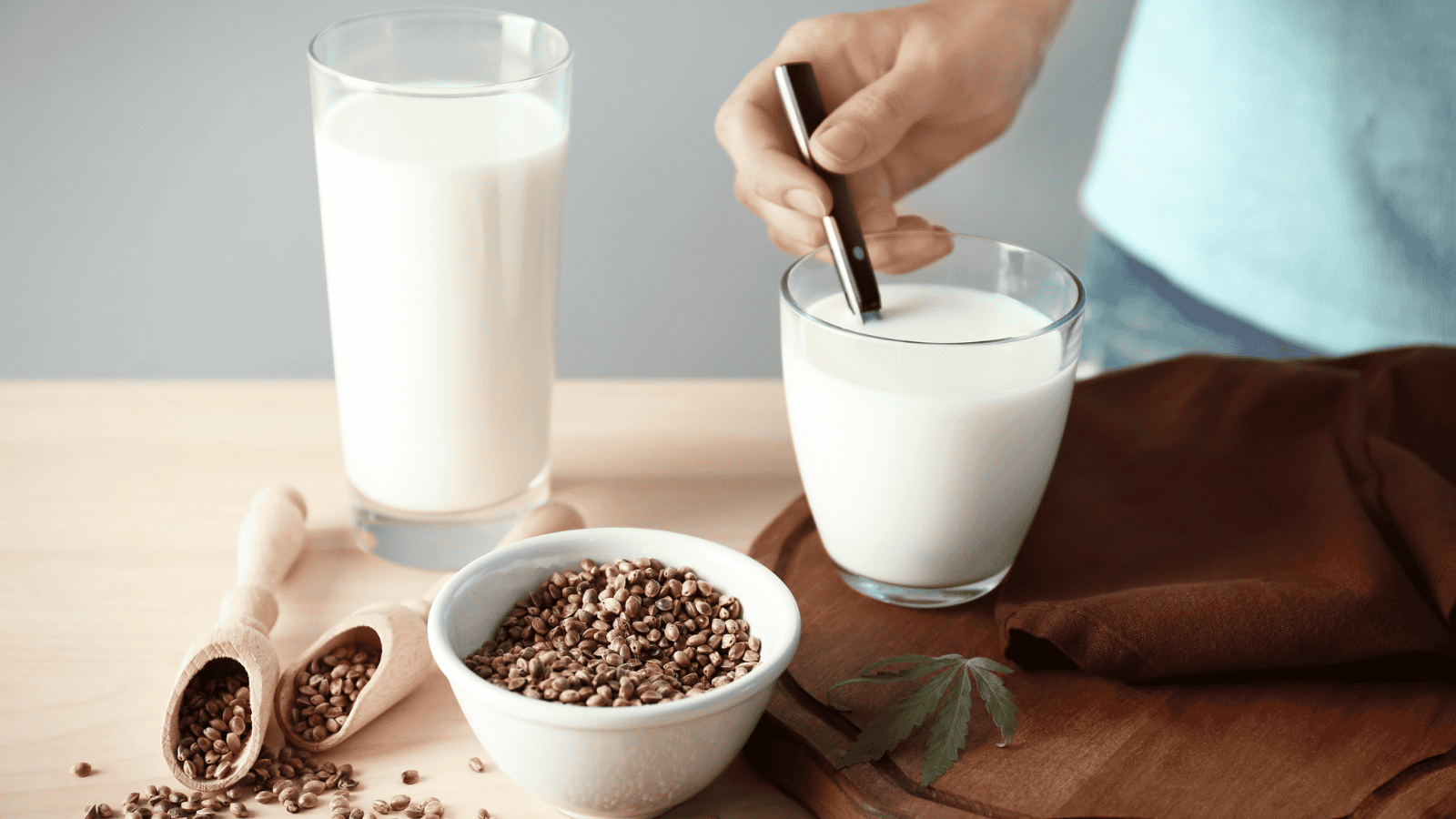 A person stirs a glass of hemp milk, a popular plant-based dairy milk substitute, with a metal straw. Another glass, a bowl of hemp seeds, and two wooden scoops filled with these nutritious seeds are on the table alongside a brown cloth.