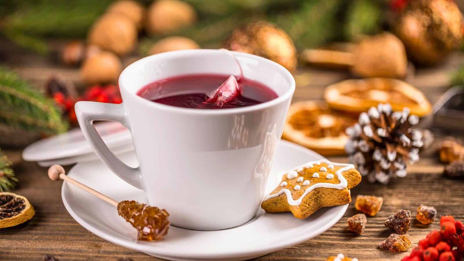 A white cup filled with a red beverage sits on a saucer. A gingerbread cookie with white icing and a crystalized sugar stick are placed on the saucer. In the background, there are Christmas decorations, dried fruits, and nuts.