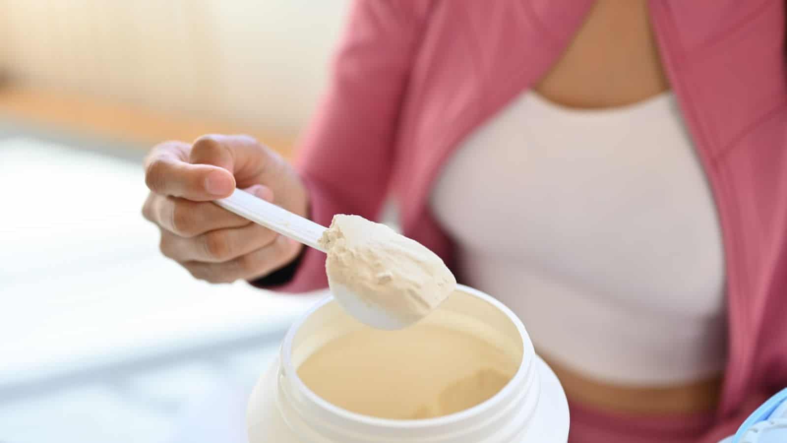 A person wearing a pink jacket and white top is scooping protein powder from a container using a white scoop. The container is white, and the focus is on the scoop and the powder. The background is blurred.