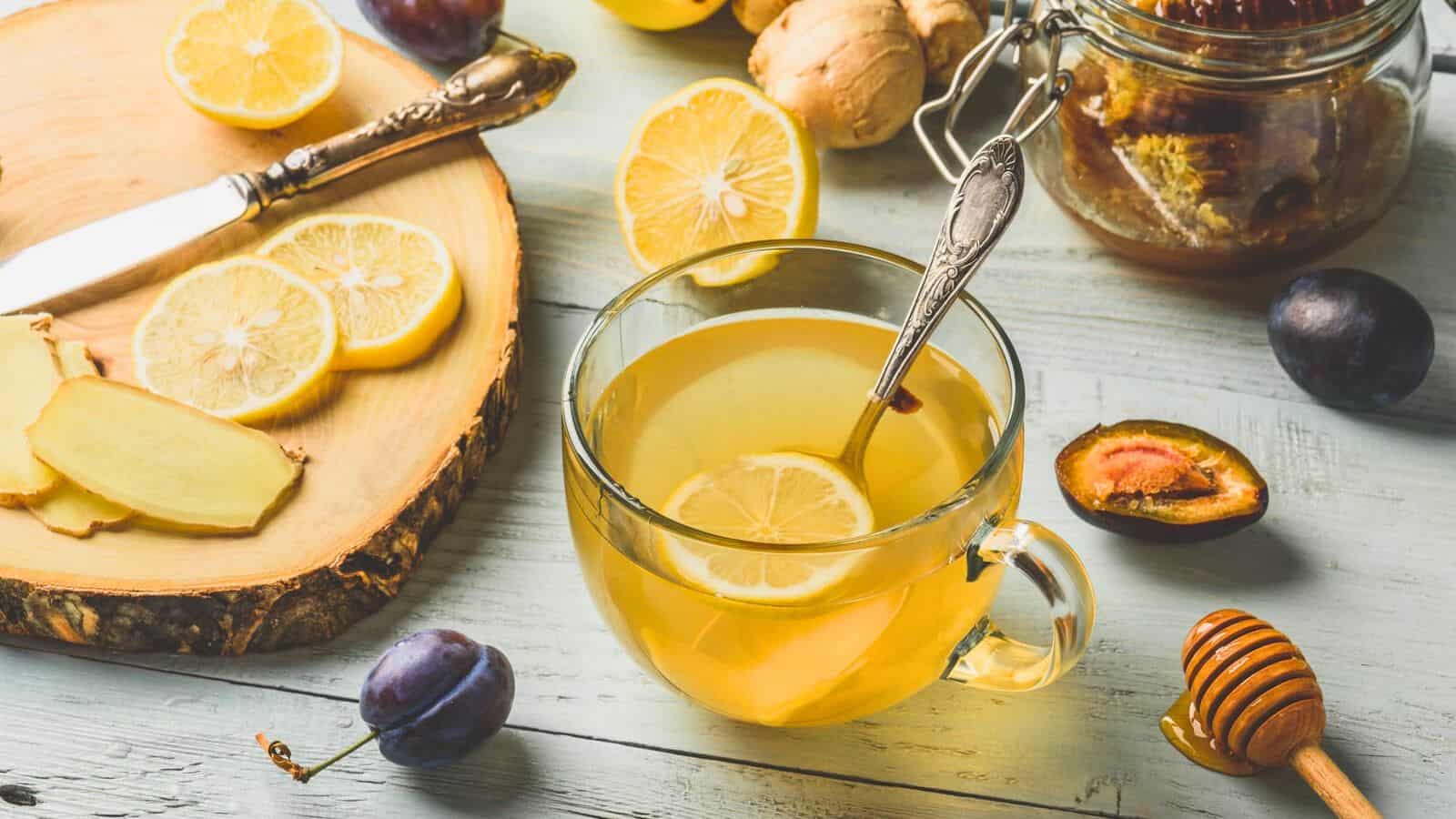 A glass mug filled with honey lemon tea, garnished with a lemon slice. Nearby are lemon slices, ginger slices, a jar of honey, halved plums, a honey dipper, and a knife on a wooden board. The items are arranged on a light-colored wooden surface.