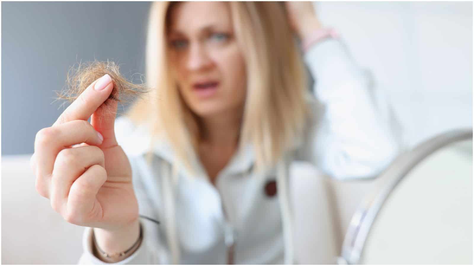 A person holds a clump of hair in their hand while looking distressed. The focus is on the hair, with the person's blurred face and hand in the background. They are wearing a light-colored top.