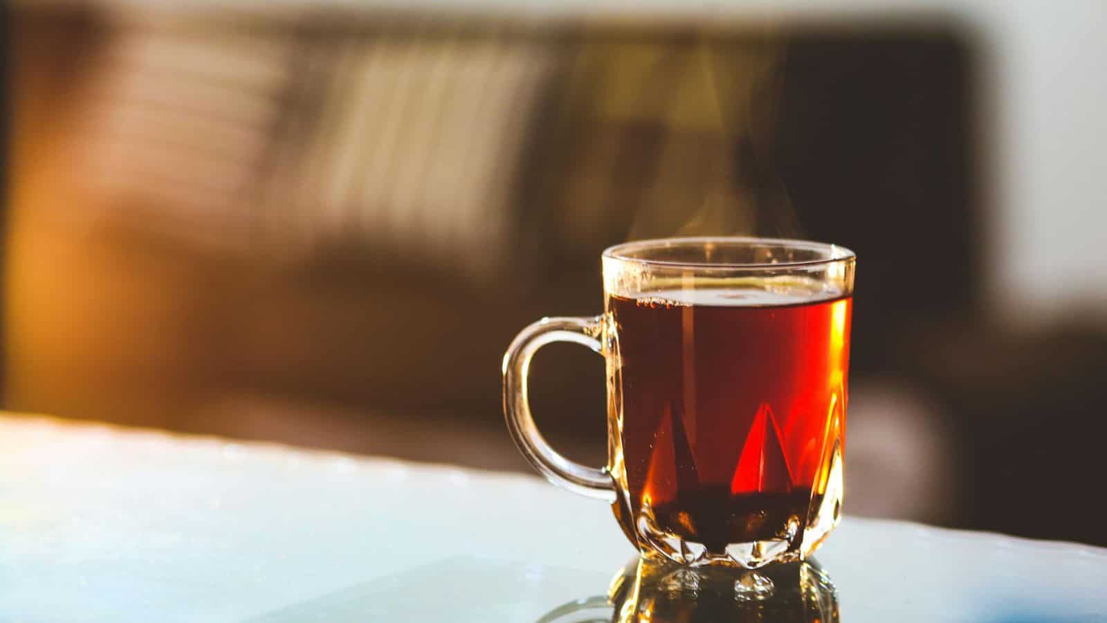 A clear glass mug filled with hot tea is placed on a reflective surface. The tea appears to be steaming. In the background, there&rsquo;s a blurred image of a sofa with cushions. Warm sunlight illuminates part of the image, creating a cozy atmosphere.