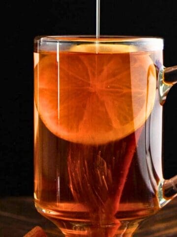 A clear glass mug filled with a dark amber liquid, garnished with a lemon slice and a cinnamon stick. A stream of honey is being drizzled the drink. The background is dark, and the mug is placed on a wooden surface.