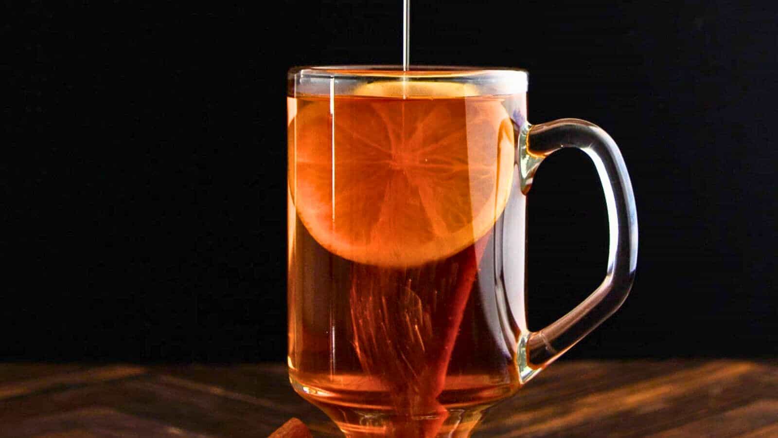 A clear glass mug filled with a dark amber liquid, garnished with a lemon slice and a cinnamon stick. A stream of honey is being drizzled the drink. The background is dark, and the mug is placed on a wooden surface.