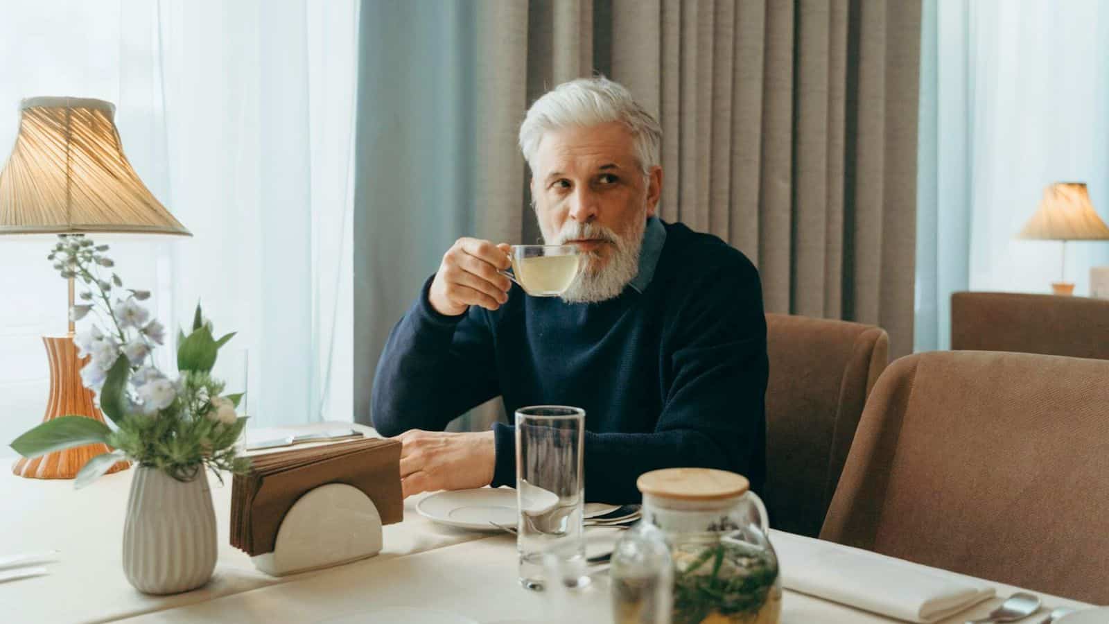 An older man with a white beard, dressed in a dark sweater, is sitting at a restaurant table. He is raising a cup of tea to his lips. The table is set with a vase of flowers, a napkin holder, glass, and a jar with green herbs.
