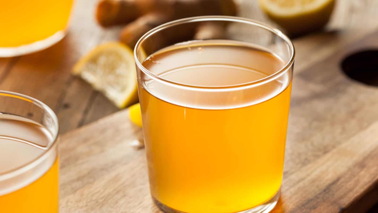 A close-up of a glass filled with kombucha. A lemon wedge and a piece of ginger are partially visible in the background.