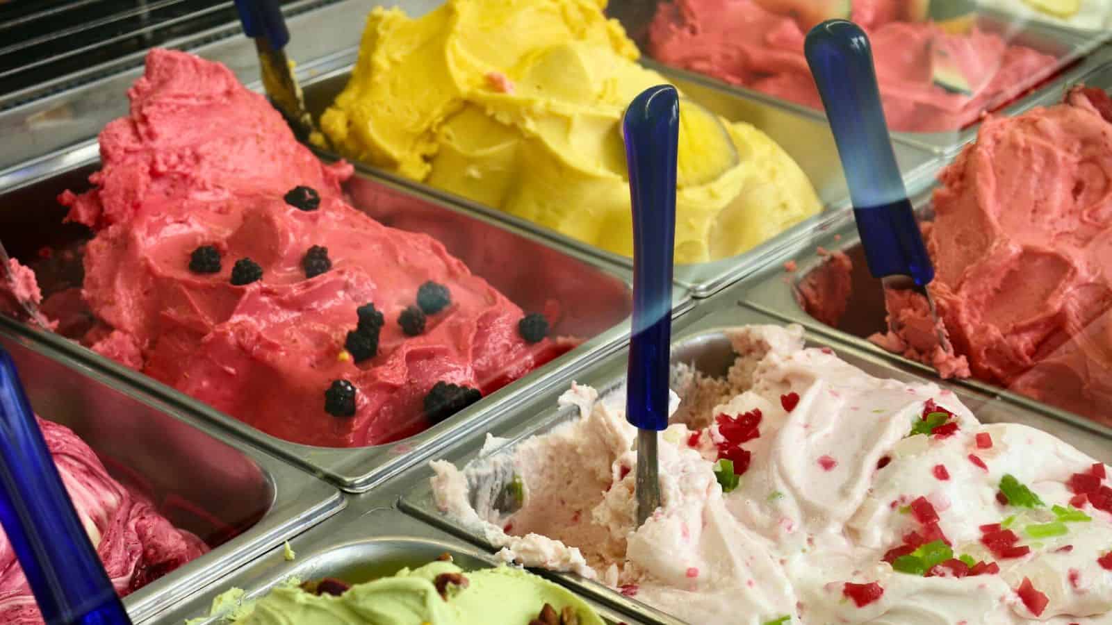 A display case filled with various flavors of ice cream or gelato in metal containers. The flavors include pink, yellow, green, and white, with some adorned with toppings such as berries and small pieces of fruit. Blue scoops are placed in each container.