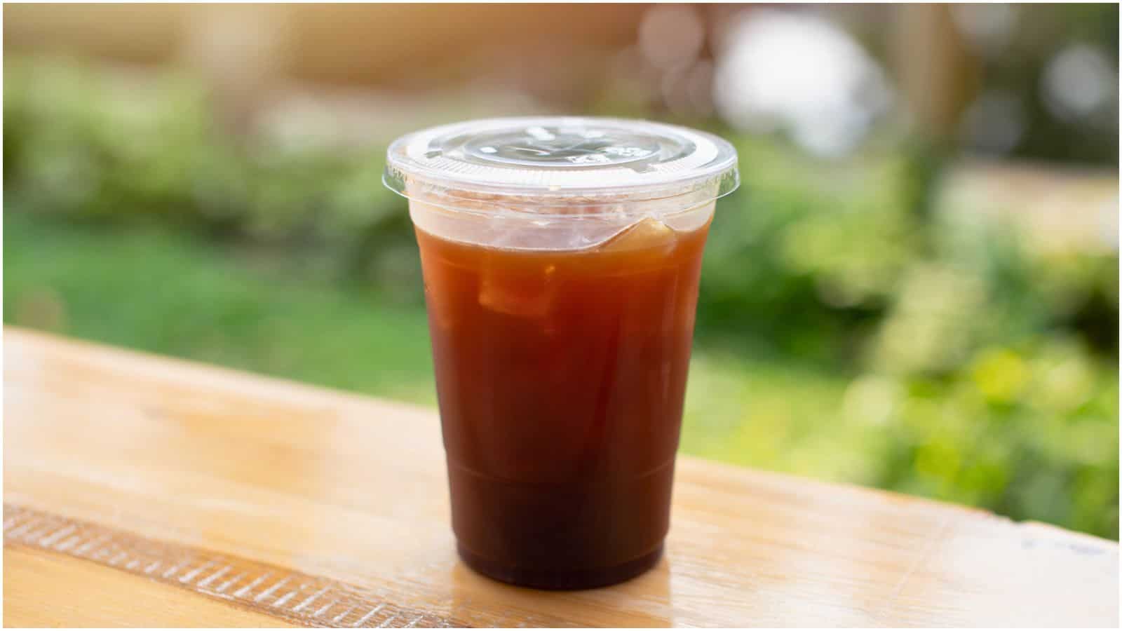 A plastic cup filled with iced coffee, covered with a clear plastic lid, sits on a wooden surface. The background is slightly blurred, showing greenery and hints of light.