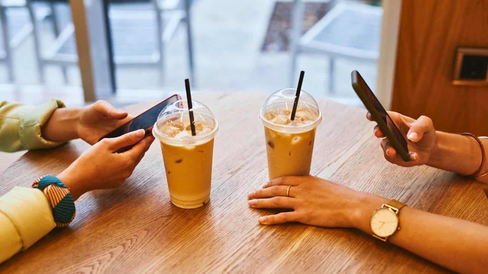 Two iced coffee drinks with clear lids and straws are on a wooden table. Two hands holding smartphones are visible, one wearing a watch and the other a bracelet. The background features blurred chairs and a window.