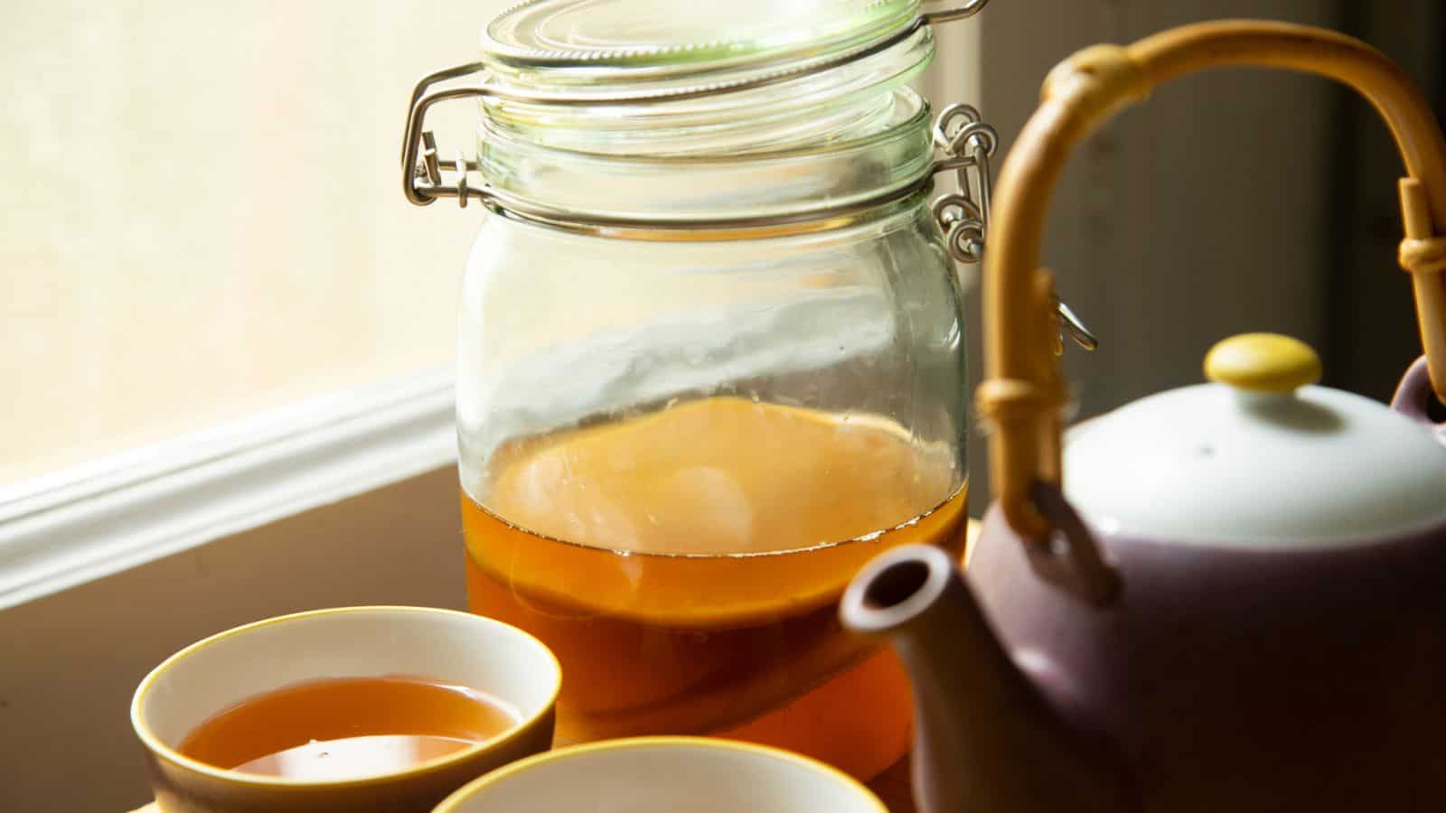 A large glass jar filled with kombucha sits on a windowsill. Beside the jar are two small teacups filled with the same beverage and a purple teapot with a bamboo handle.