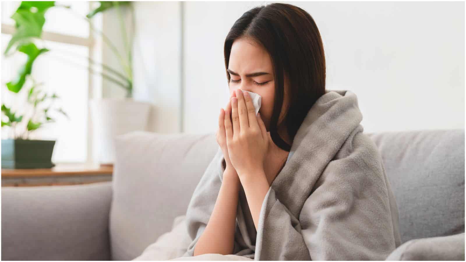 A woman is sitting on a couch, wrapped in a grey blanket, and blowing her nose with a tissue. A potted plant is visible in the background. The setting suggests she might be unwell.