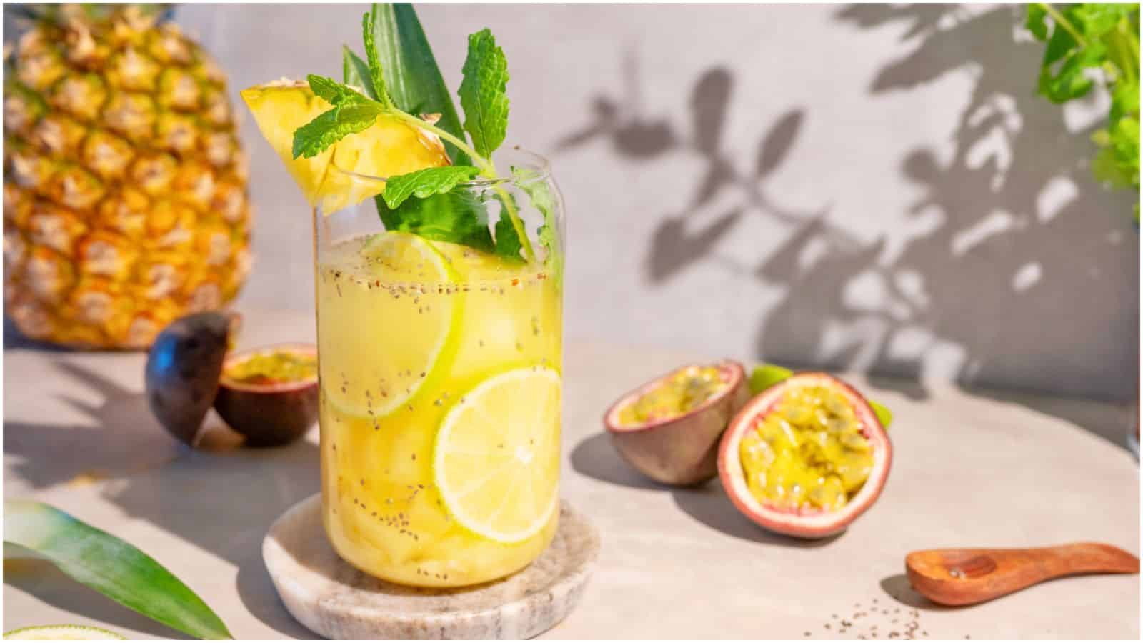 A glass of pineapple and passion fruit drink garnished with a slice of pineapple, lemon, and fresh mint leaves sits on a coaster. With half-cut passion fruits and a whole pineapple in the background on a sunlit surface.