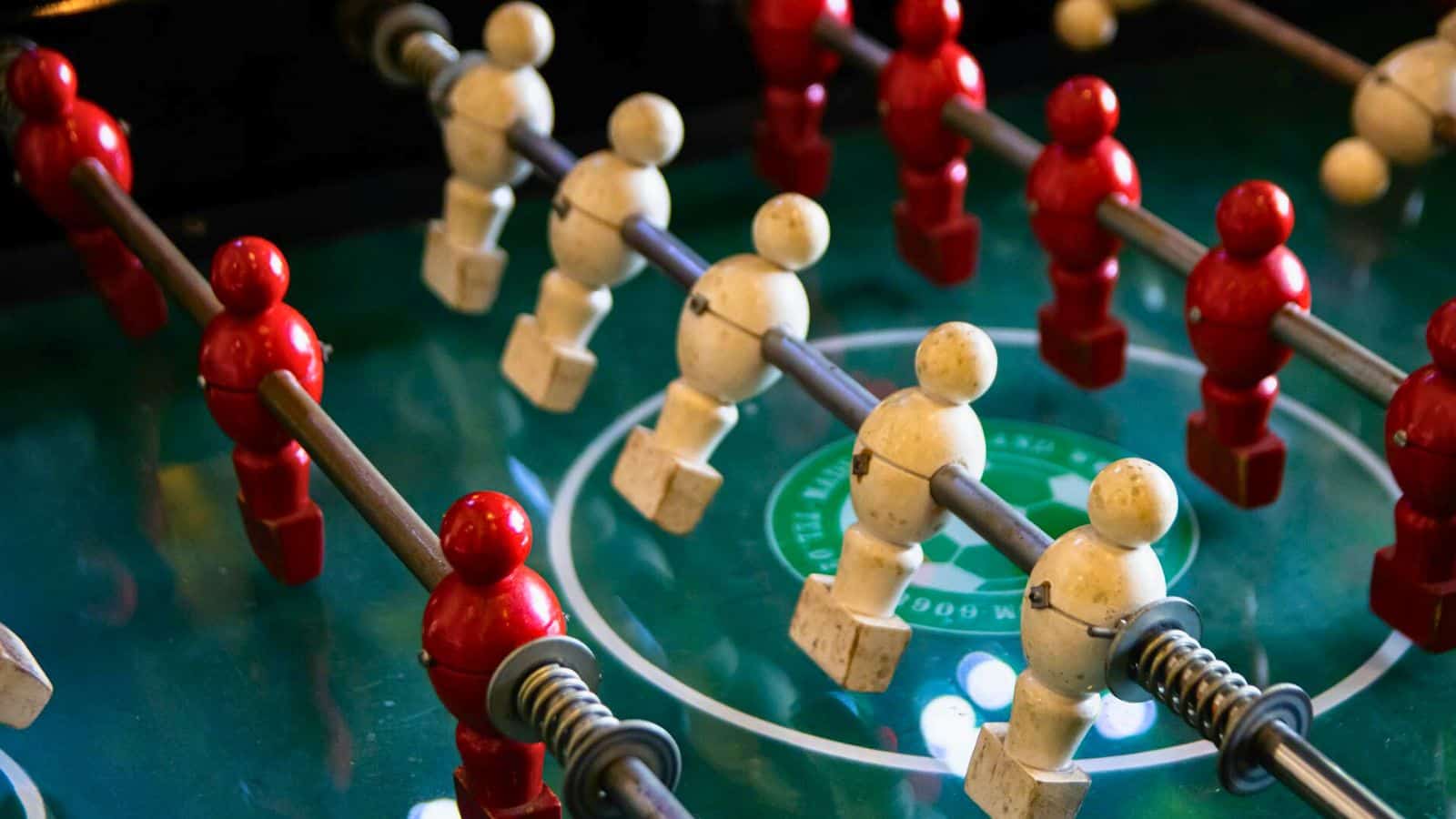 A close-up view of a foosball table. The table has alternating rows of red and white player figures, mounted on metal rods. The surface is green with markings, and the figures are in a stationary position.