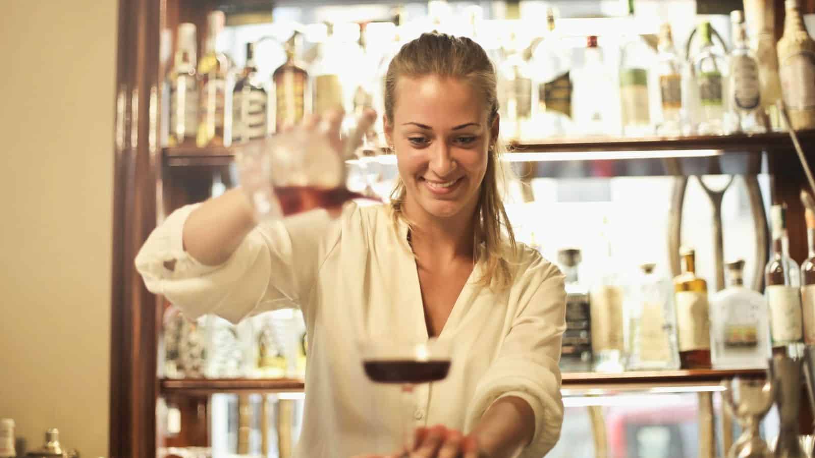 A bartender smiles while pouring a fun drink behind a bar counter. Shelves filled with various bottles of alcohol are visible in the background. She wears a long-sleeved white shirt and uses two hands to pour from a shaker into a glass.