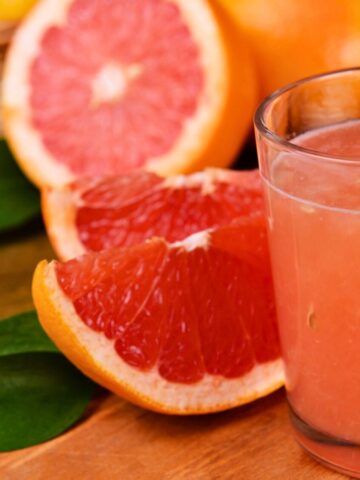 A close-up photo of a glass filled with pink grapefruit juice placed on a wooden surface. Fresh grapefruit slices and whole grapefruits are in the background. A basket of fruits and green leaves are also visible.