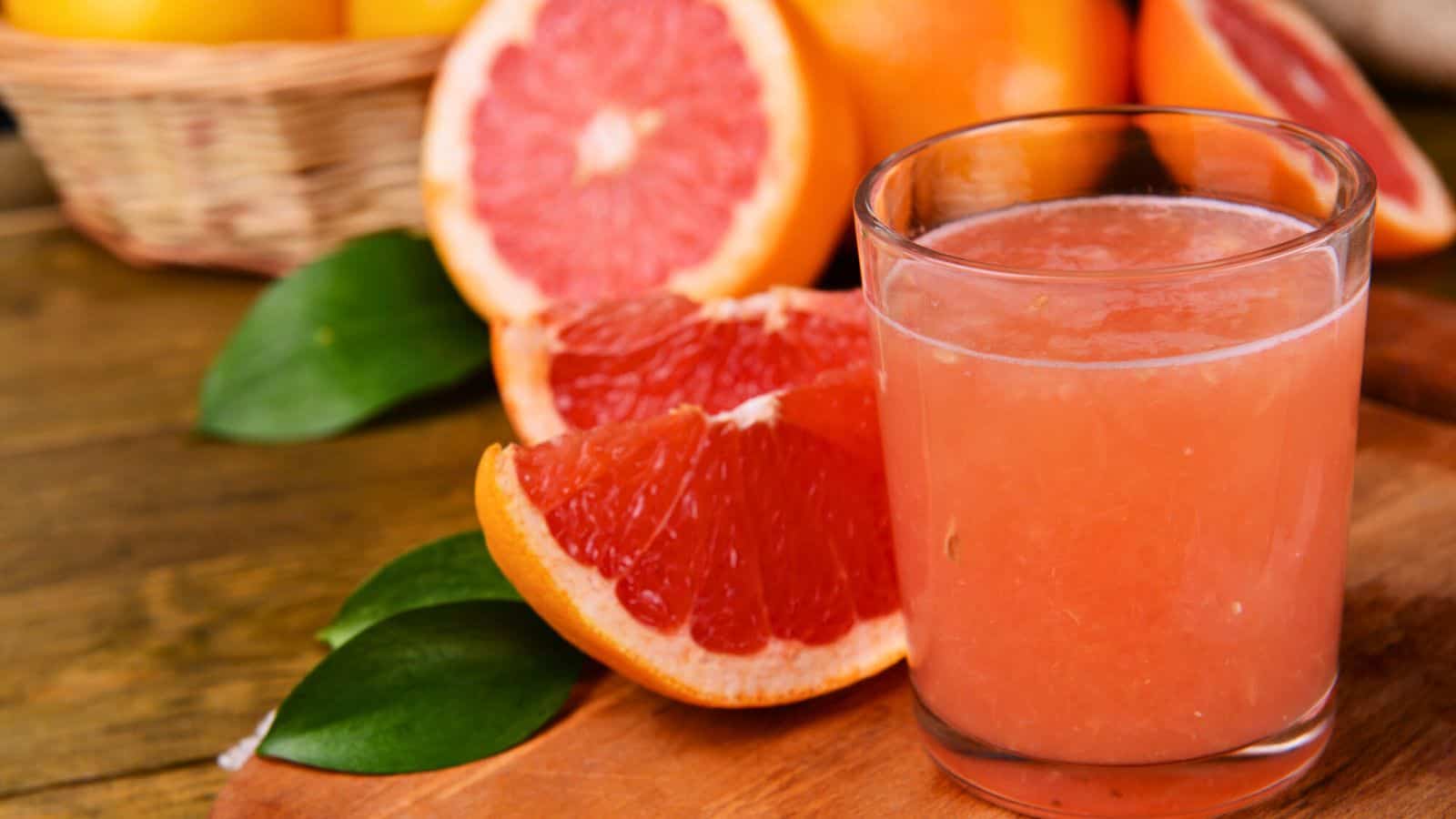 A close-up photo of a glass filled with pink grapefruit juice placed on a wooden surface. Fresh grapefruit slices and whole grapefruits are in the background. A basket of fruits and green leaves are also visible.