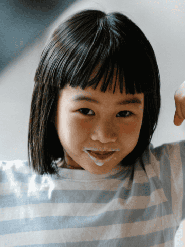 A child with a milk mustache holds a glass of plant-based dairy milk in one hand and flexes their other arm in a playful pose. They are wearing a striped shirt and a wristwatch.