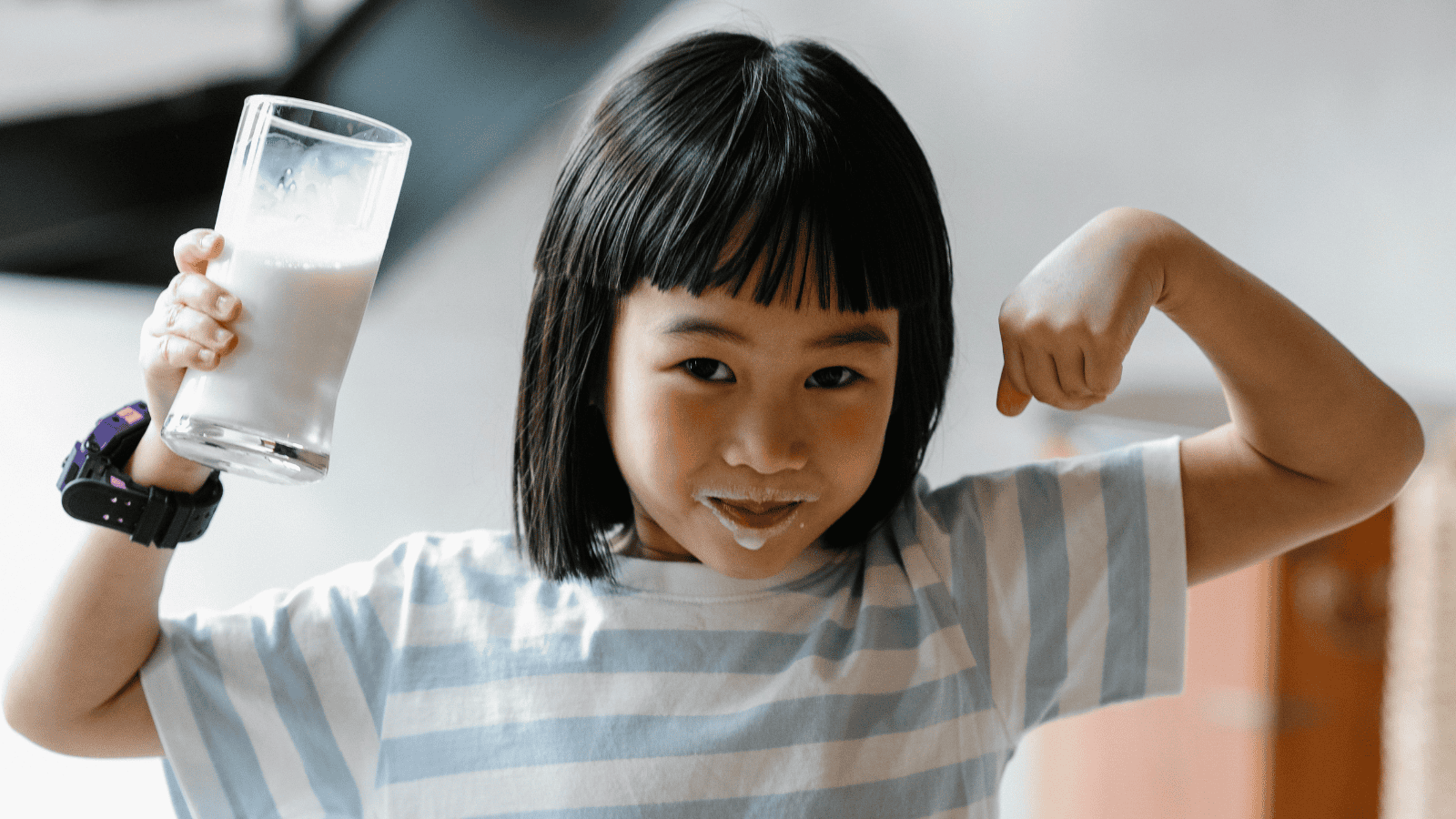 A child with a milk mustache holds a glass of plant-based dairy milk in one hand and flexes their other arm in a playful pose. They are wearing a striped shirt and a wristwatch.