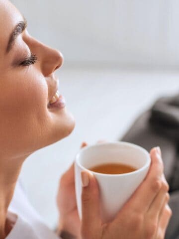 A woman with brown hair is sitting indoors, holding a white mug close to her face. Her eyes are closed, and she has a content expression. There is a couch with pillows in the blurred background.
