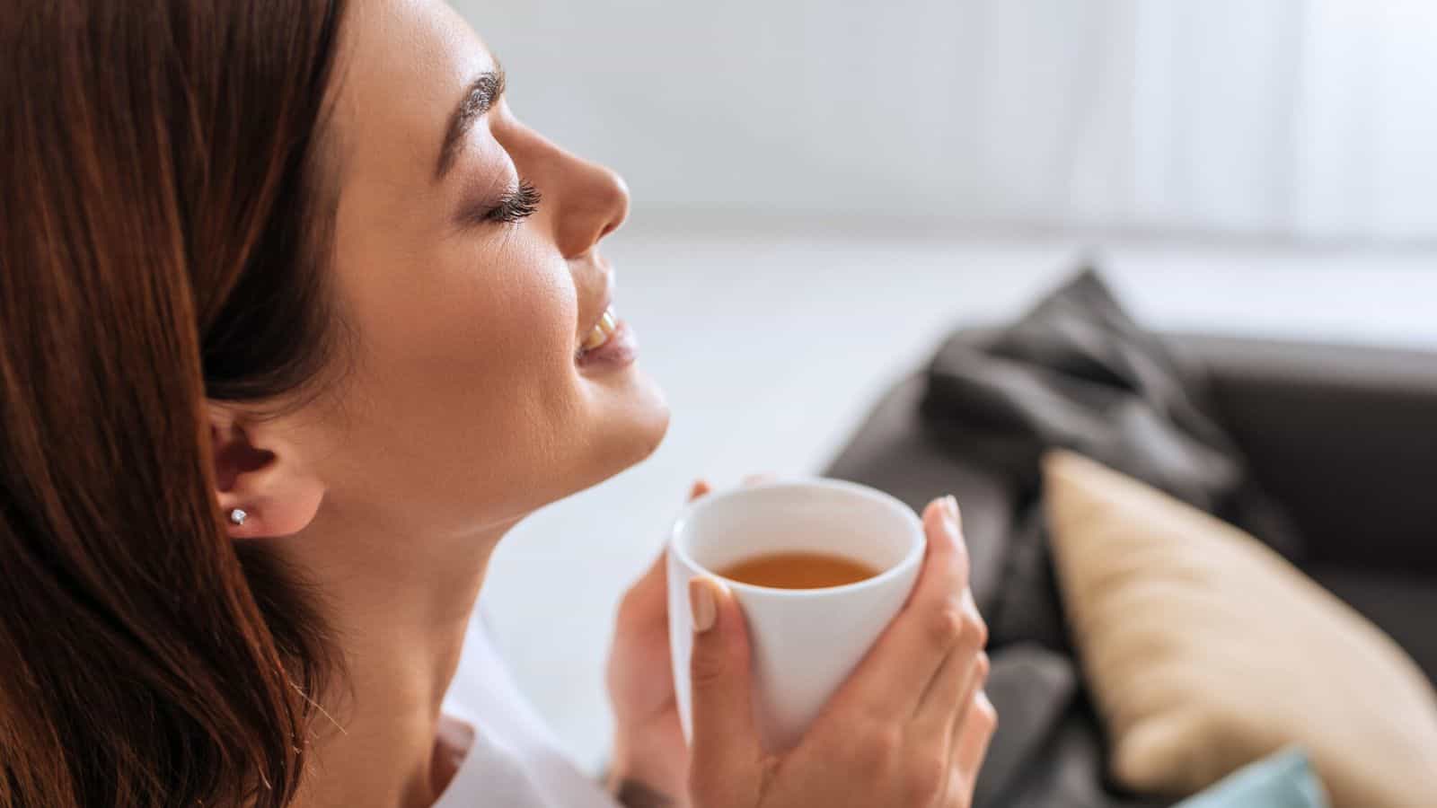A woman with brown hair is sitting indoors, holding a white mug close to her face. Her eyes are closed, and she has a content expression. There is a couch with pillows in the blurred background.