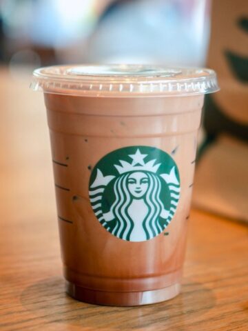 A small, clear plastic cup with a lid, containing an iced coffee beverage, sits on a wooden table. The cup features the Starbucks logo. In the background, there is a blurry Starbucks paper bag and a partially visible Starbucks packet on the right side of the table.