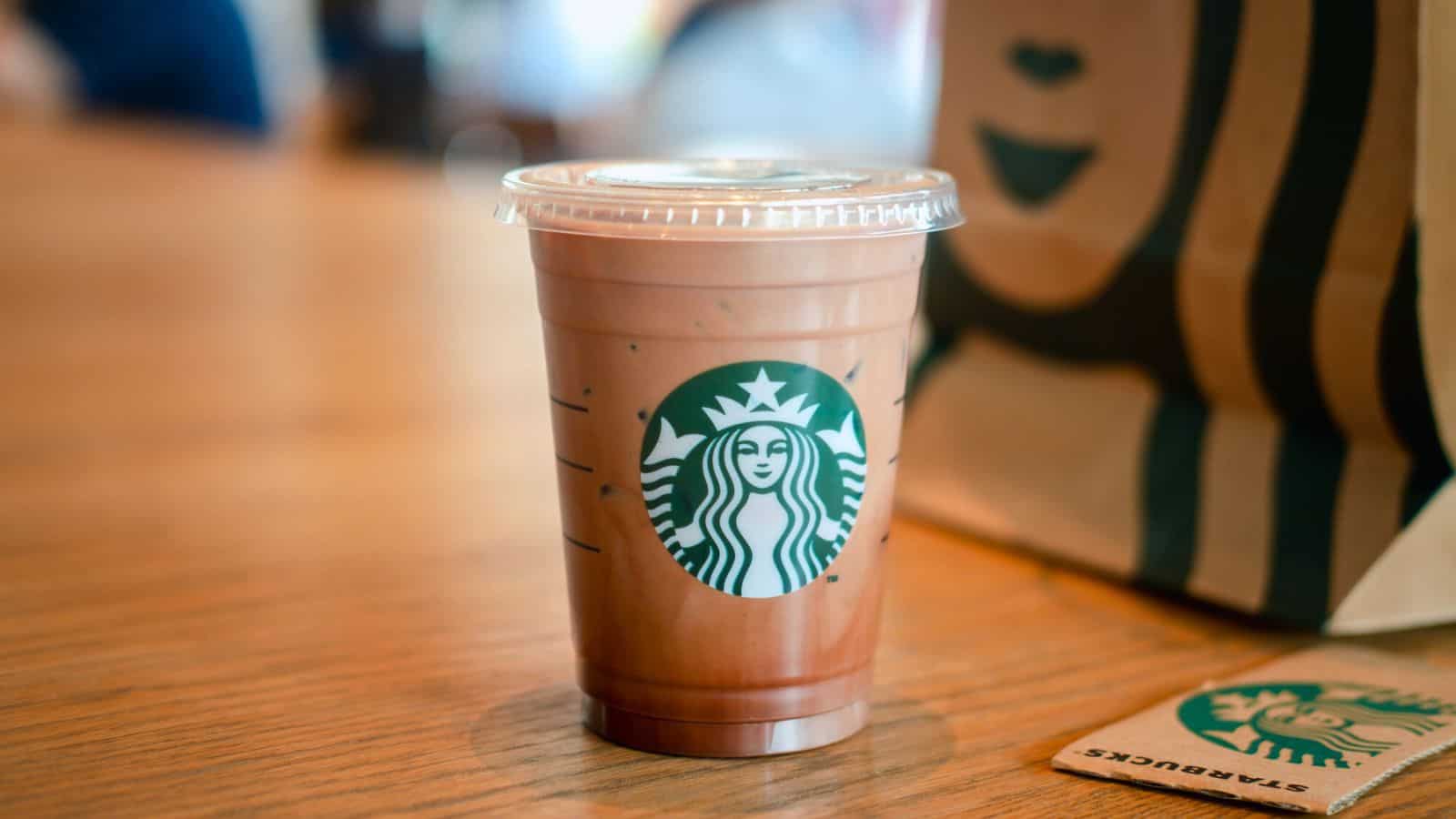 A small, clear plastic cup with a lid, containing an iced coffee beverage, sits on a wooden table. The cup features the Starbucks logo. In the background, there is a blurry Starbucks paper bag and a partially visible Starbucks packet on the right side of the table.