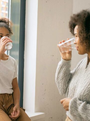 Two people are drinking water. One person, standing, wears a grey sweater and jeans. The other, sitting on a window ledge in a white t-shirt and brown pants. They are indoors with a large window showcasing an urban scene outside.
