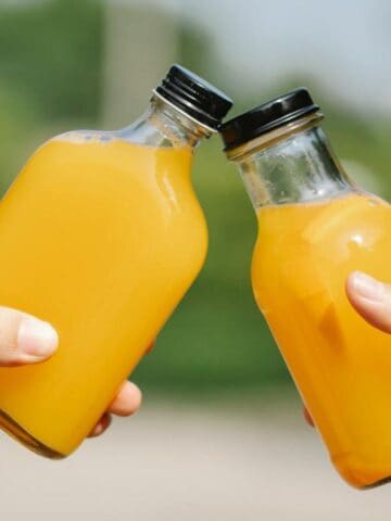 Two hands holding small glass bottles of orange juice, clinking them together in a toasting gesture. The background is blurred greenery.
