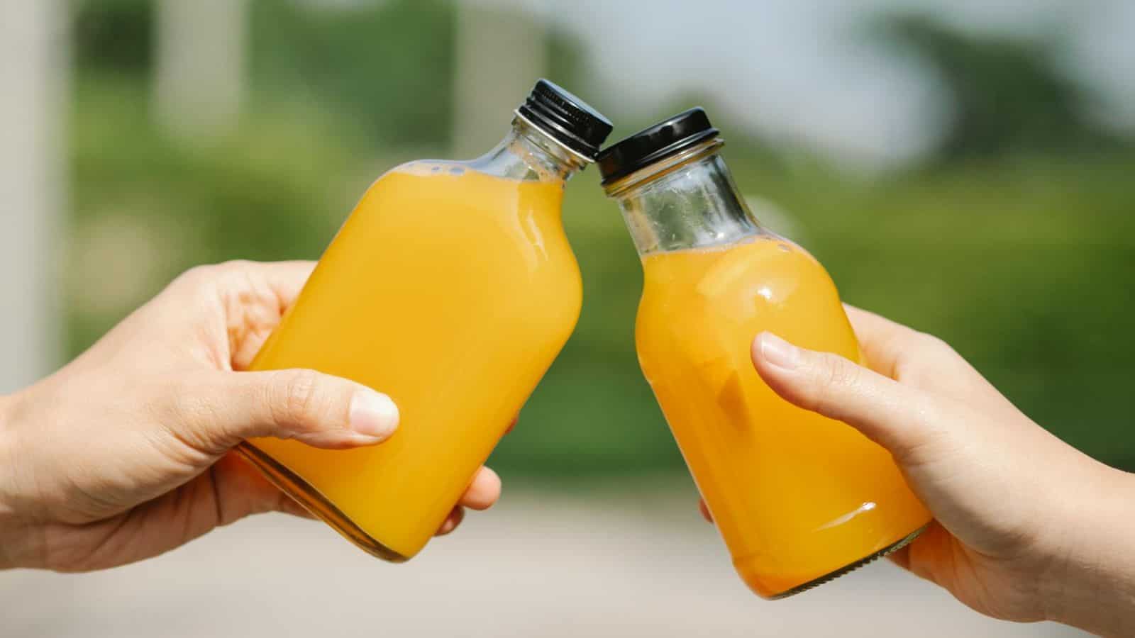 Two hands holding small glass bottles of orange juice, clinking them together in a toasting gesture. The background is blurred greenery.