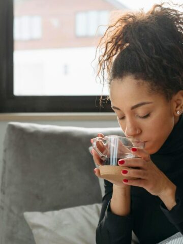 A woman with curly hair is sitting on a couch, holding a clear glass mug with both hands and drinking from it. She is wearing a black turtleneck sweater, and behind her is a large window with a view of buildings in the distance.
