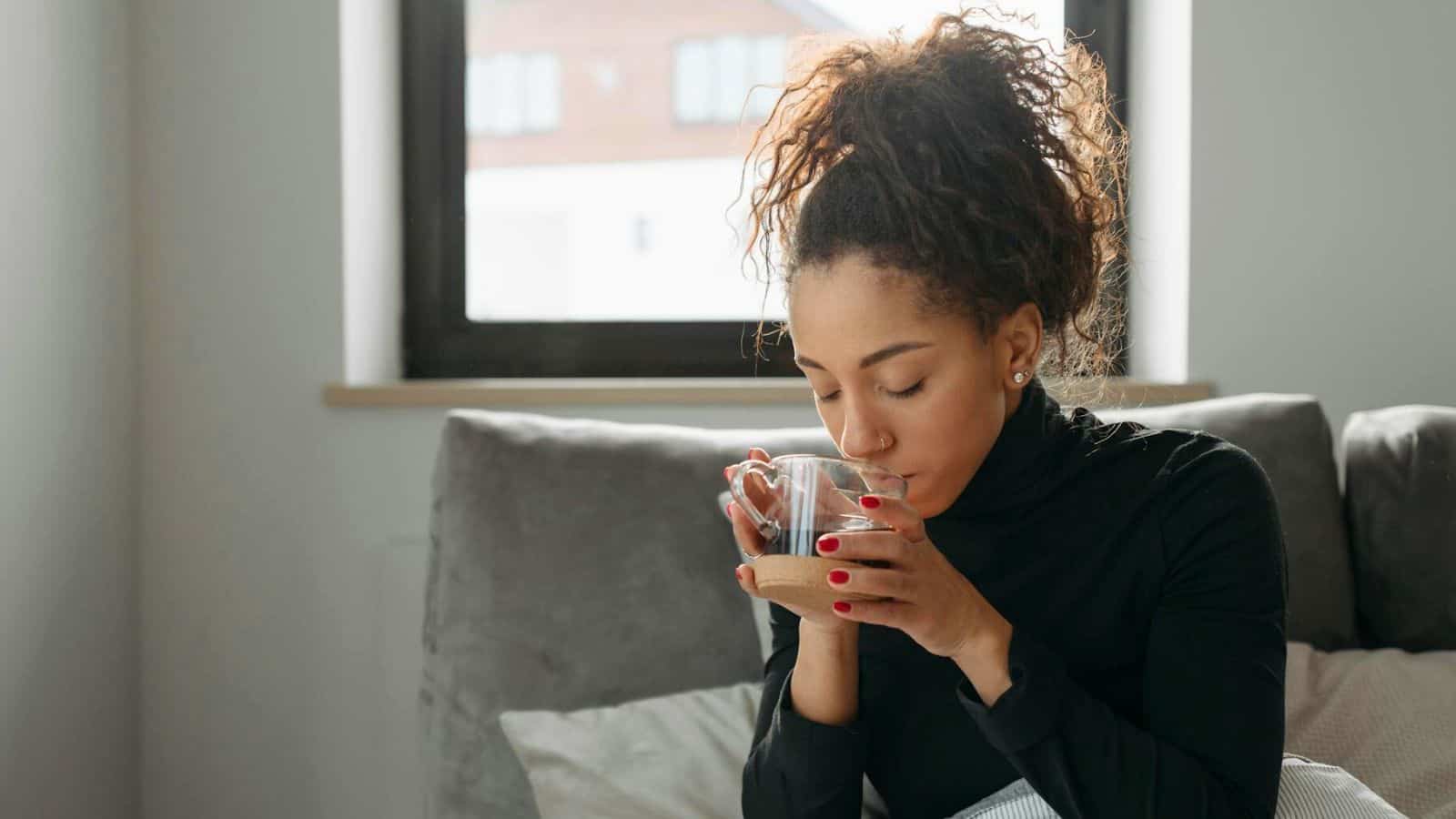 A woman with curly hair is sitting on a couch, holding a clear glass mug with both hands and drinking from it. She is wearing a black turtleneck sweater, and behind her is a large window with a view of buildings in the distance. 