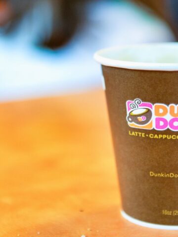 A Dunkin' Donuts cup filled with a hot beverage sits on a table. The cup features the Dunkin' Donuts logo and the text "Latte, Cappuccino, Espresso." A blurred background and an out-of-focus person can also be seen.