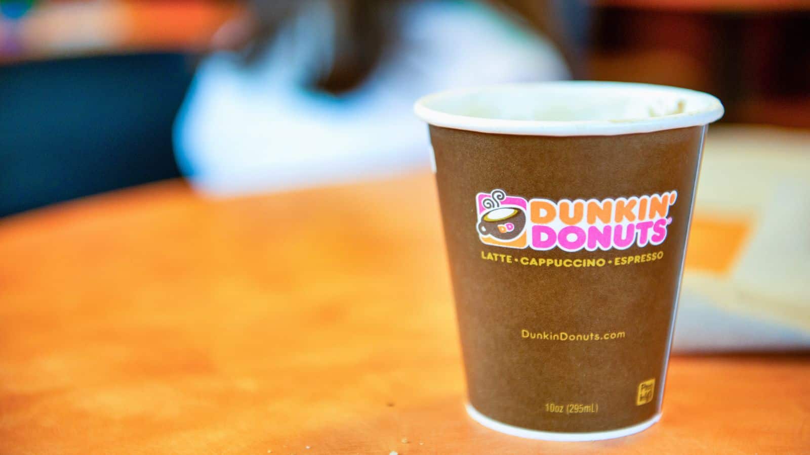 A Dunkin' Donuts cup filled with a hot beverage sits on a table. The cup features the Dunkin' Donuts logo and the text "Latte, Cappuccino, Espresso." A blurred background and an out-of-focus person can also be seen.