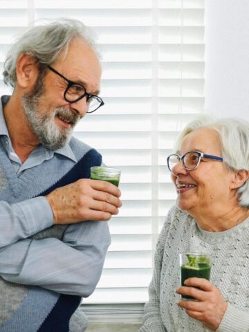An elderly man and woman are smiling at each other while holding green smoothies. The man is wearing a light blue shirt with a gray argyle sweater vest, and the woman is in a gray knit sweater. They are standing in front of white blinds.