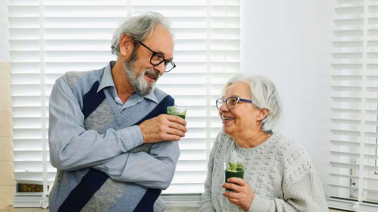 An elderly man and woman are smiling at each other while holding green smoothies. The man is wearing a light blue shirt with a gray argyle sweater vest, and the woman is in a gray knit sweater. They are standing in front of white blinds.