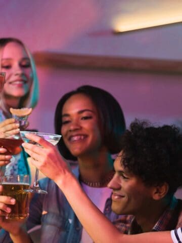 Five people are gathered in a celebratory atmosphere, raising glasses in a toast. They are smiling and appear to be enjoying each other's company. The background has bright ambient lighting, and the group is holding various types of beverages.