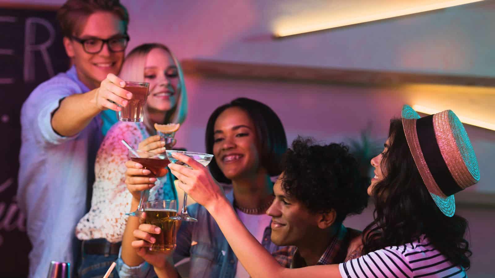 Five people are gathered in a celebratory atmosphere, raising glasses in a toast. They are smiling and appear to be enjoying each other's company. The background has bright ambient lighting, and the group is holding various types of beverages.