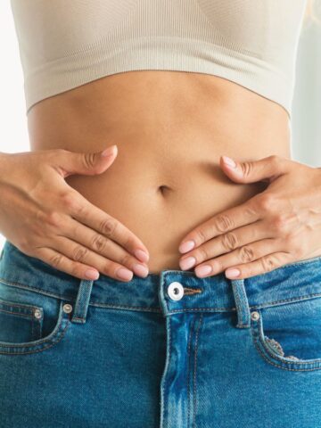A person in a cropped white top and blue jeans is standing while placing their hands on their stomach. The background is blurred.