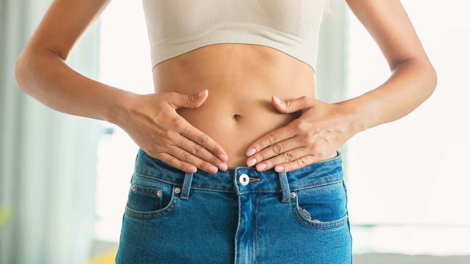A person in a cropped white top and blue jeans is standing while placing their hands on their stomach. The background is blurred.