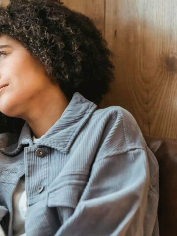 A person with curly hair sits on a brown sofa, holding a tissue to their face. They appear to be looking out the window with a thoughtful or sad expression. The background features a wooden wall.