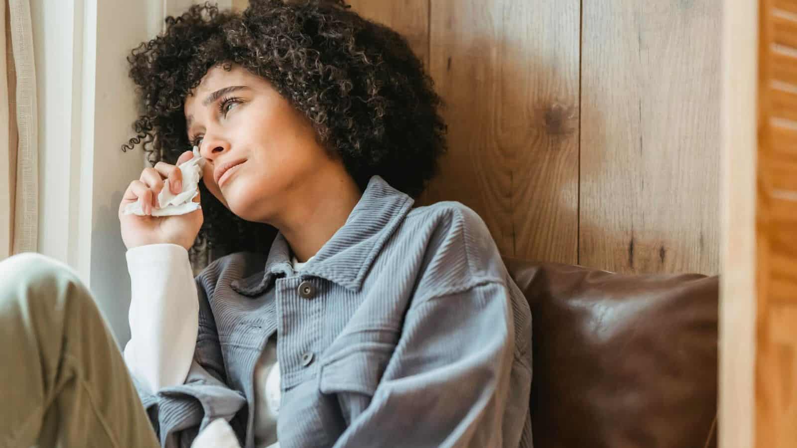 A person with curly hair sits on a brown sofa, holding a tissue to their face. They appear to be looking out the window with a thoughtful or sad expression. The background features a wooden wall.