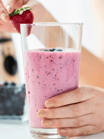 A person holds a glass of superfood smoothie with blueberries, placing a strawberry on the rim. A blender, a bowl of strawberries, and blueberries grace the table. The background is softly blurred, highlighting this nutritious delight.