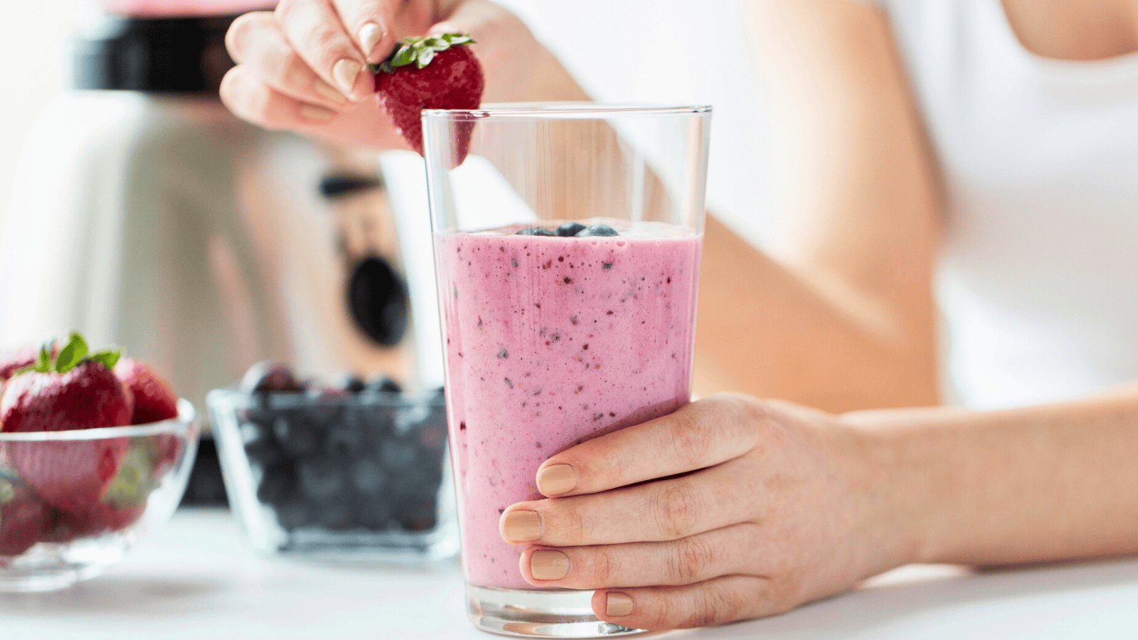 A person holds a glass of superfood smoothie with blueberries, placing a strawberry on the rim. A blender, a bowl of strawberries, and blueberries grace the table. The background is softly blurred, highlighting this nutritious delight.