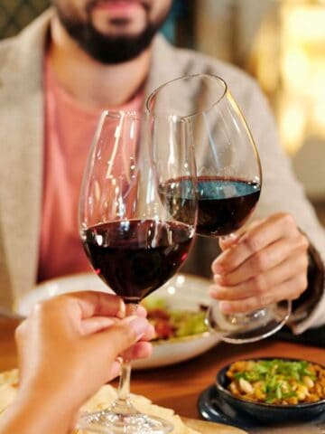 Three people clink glasses of red wine together over a table with various dishes. The background is blurred, and a vase with pink flowers is visible on the left side. The focus is on the wine glasses.