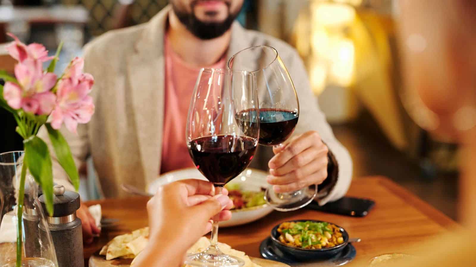 Three people clink glasses of red wine together over a table with various dishes. The background is blurred, and a vase with pink flowers is visible on the left side. The focus is on the wine glasses.