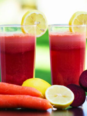 Two glasses of red juice garnished with lemon slices are placed on a table. In front of the glasses are three beetroots, two carrots, and two whole lemons, with one lemon cut in half. The background is green and out of focus.