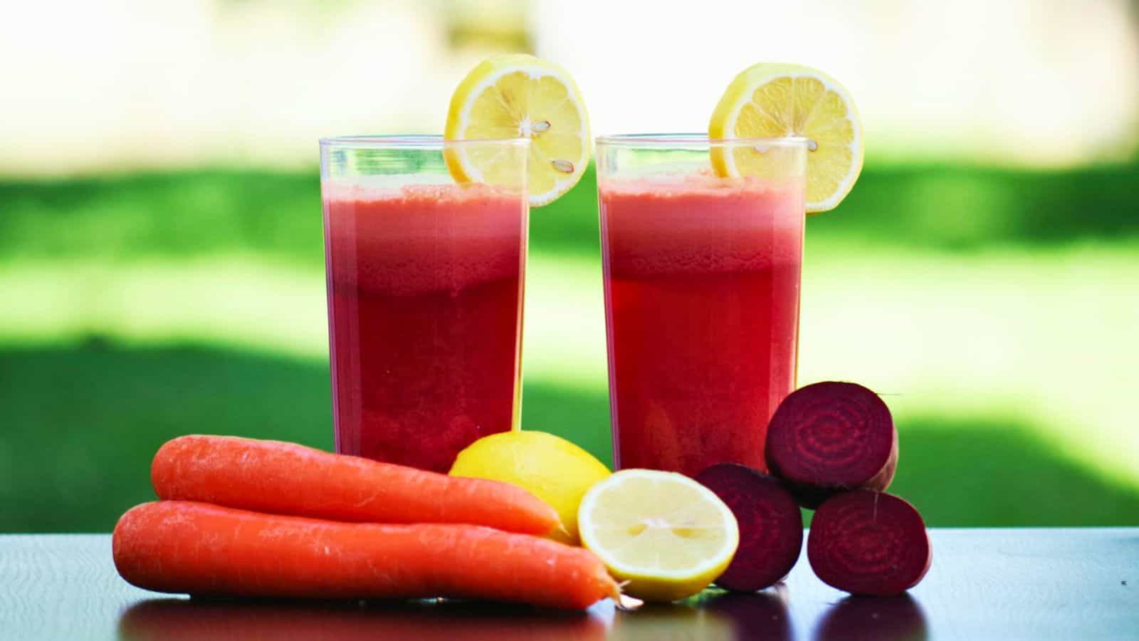 Two glasses of red juice garnished with lemon slices are placed on a table. In front of the glasses are three beetroots, two carrots, and two whole lemons, with one lemon cut in half. The background is green and out of focus.