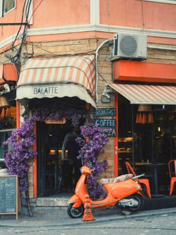 A corner café with an awning labeled "Balatte," featuring orange chairs and tables on the sidewalk. A bright orange scooter is parked in front, next to a purple-flowered vine. A small food cart or vehicle is passing by on the right side of the image.