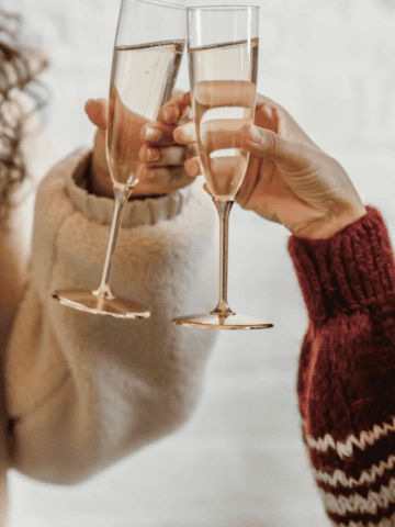 Two people clink champagne glasses together, toasting perhaps to the new harvest of unusual plants for winemaking. One wears a cream sweater, the other a striped sweater. The background is softly out of focus, suggesting an indoor celebratory setting.