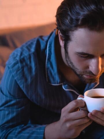 A man wearing blue pajamas is sitting on the edge of a bed, holding a white cup of tea or coffee with both hands. He is leaning forward and looking down at the cup.